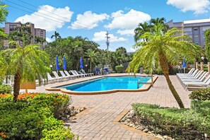 A swimming pool with lounge chairs and umbrellas, surrounded by palm trees and a brick walkway. - Breezy Condo | Deluxe Pool & Private Beach Access (Marco Island)