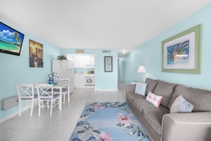 Una sala de estar con un sofá gris, una mesa de comedor con sillas, una cocina al fondo y una alfombra con motivos tropicales.