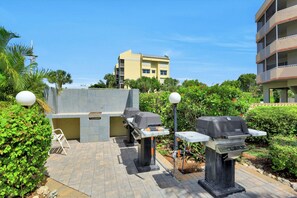 An outdoor patio area with two barbecue grills, a seating area, and a view of a yellow building in the background.