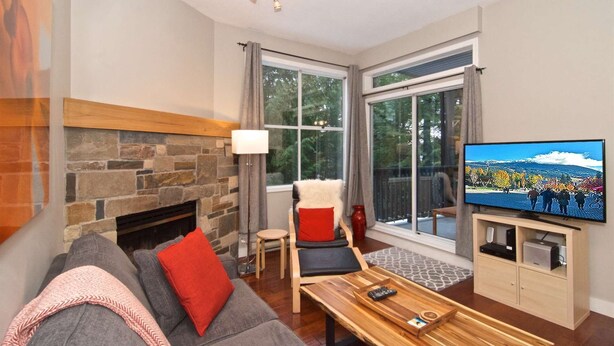 A living room with a stone fireplace, a grey sofa with red and black cushions, a wooden coffee table, and a television on a wooden cabinet. - Charming Creekside Townhouse. Easy WALK to the Gondola. Professionally Managed + Cleaned (Whistler)