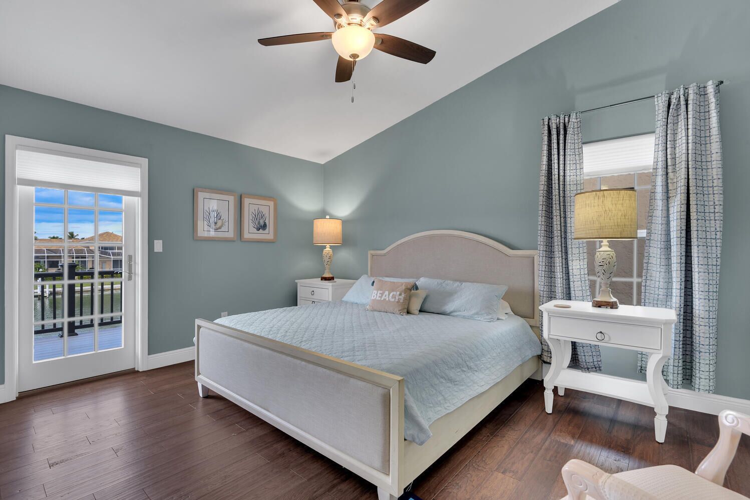 Une chambre à coucher avec un lit, un ventilateur de plafond, une table de chevet avec une lampe, une commode et une vue sur l’extérieur, visible à travers une porte.