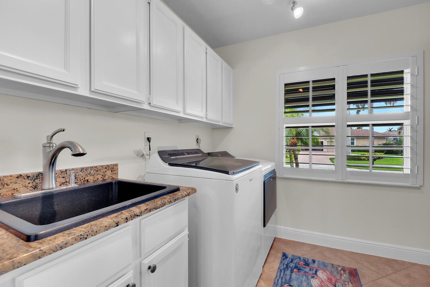 A kitchen with white cabinets, a granite countertop, a stainless steel sink, a washing machine, and a window with blinds.