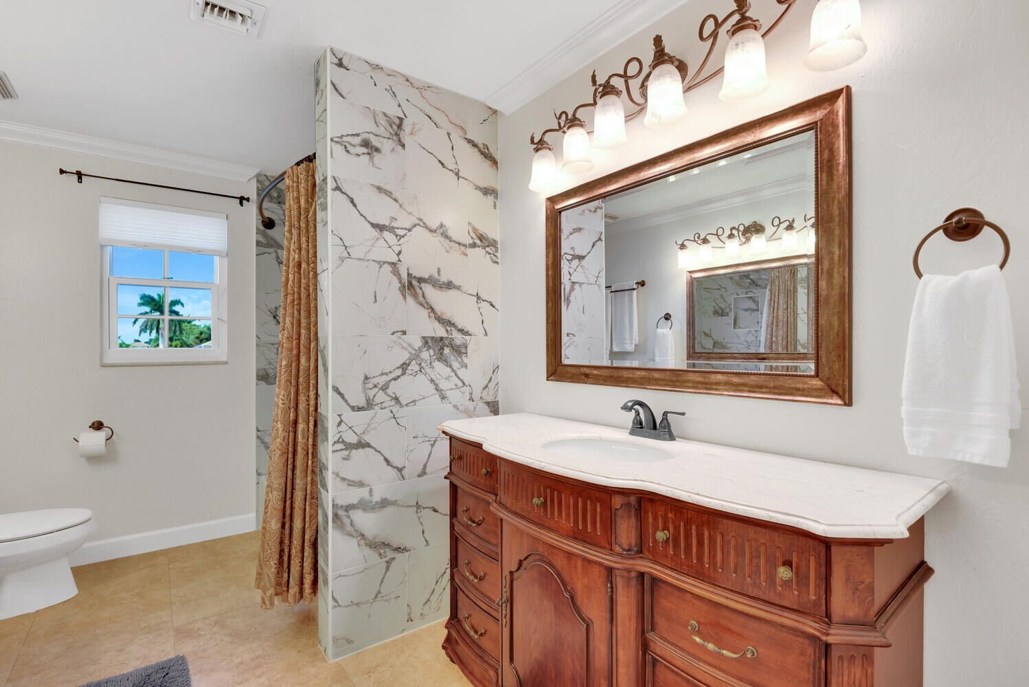 A bathroom with a marble wall, a wooden vanity, a large mirror, and a window with a view of palm trees.
