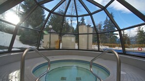 Uma área de piscina com estrutura de vidro, deck revestido de azulejos e vista para um edifício com chaminé.