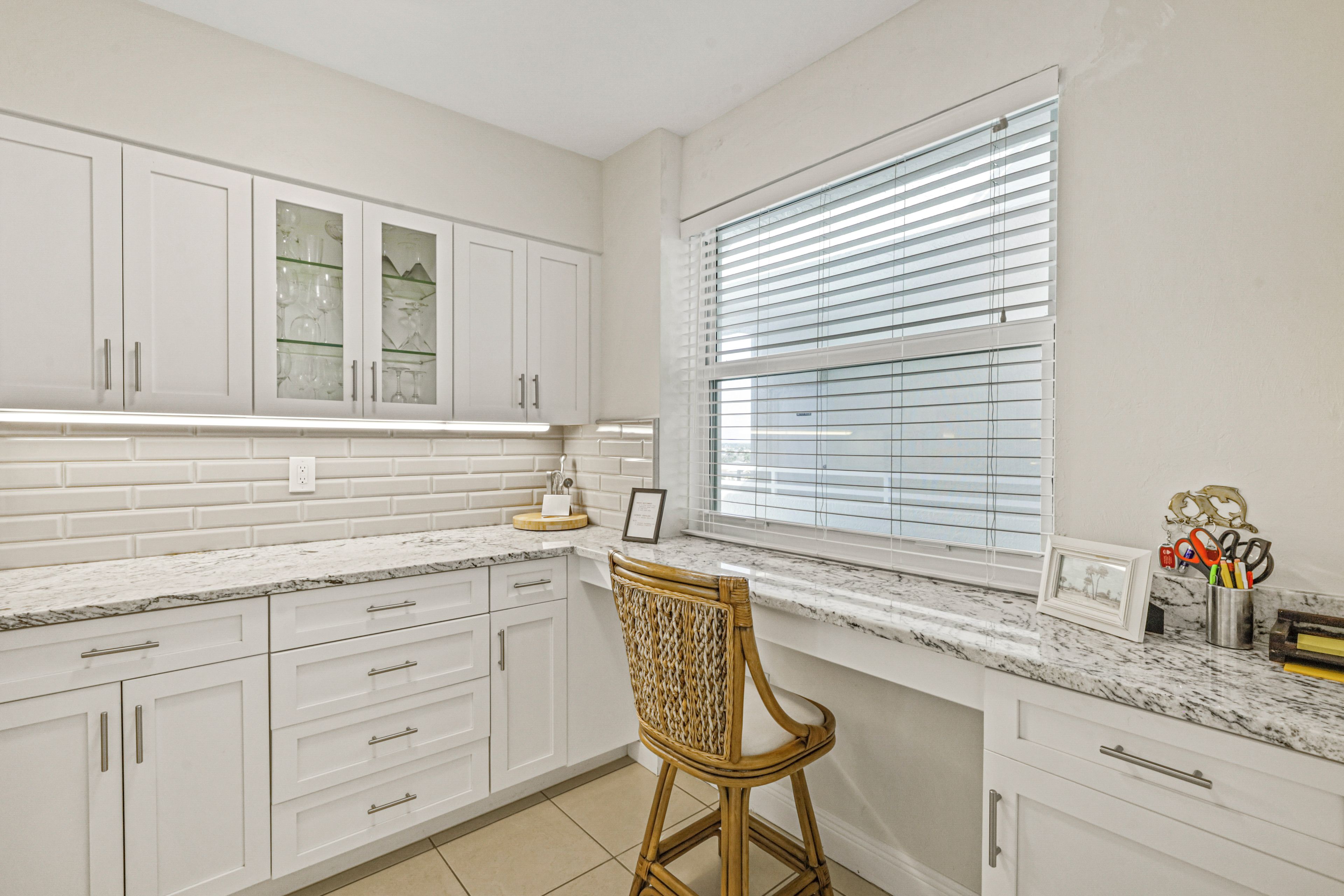 A kitchen with white cabinets, a marble countertop, a wicker chair, and a window with blinds.