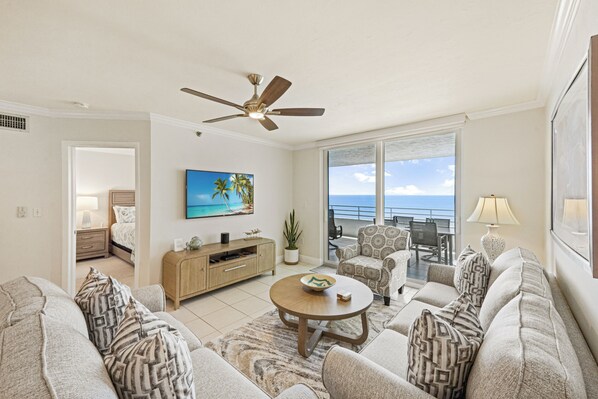 A living room with a ceiling fan, a round coffee table, and a large window with a view of the ocean.