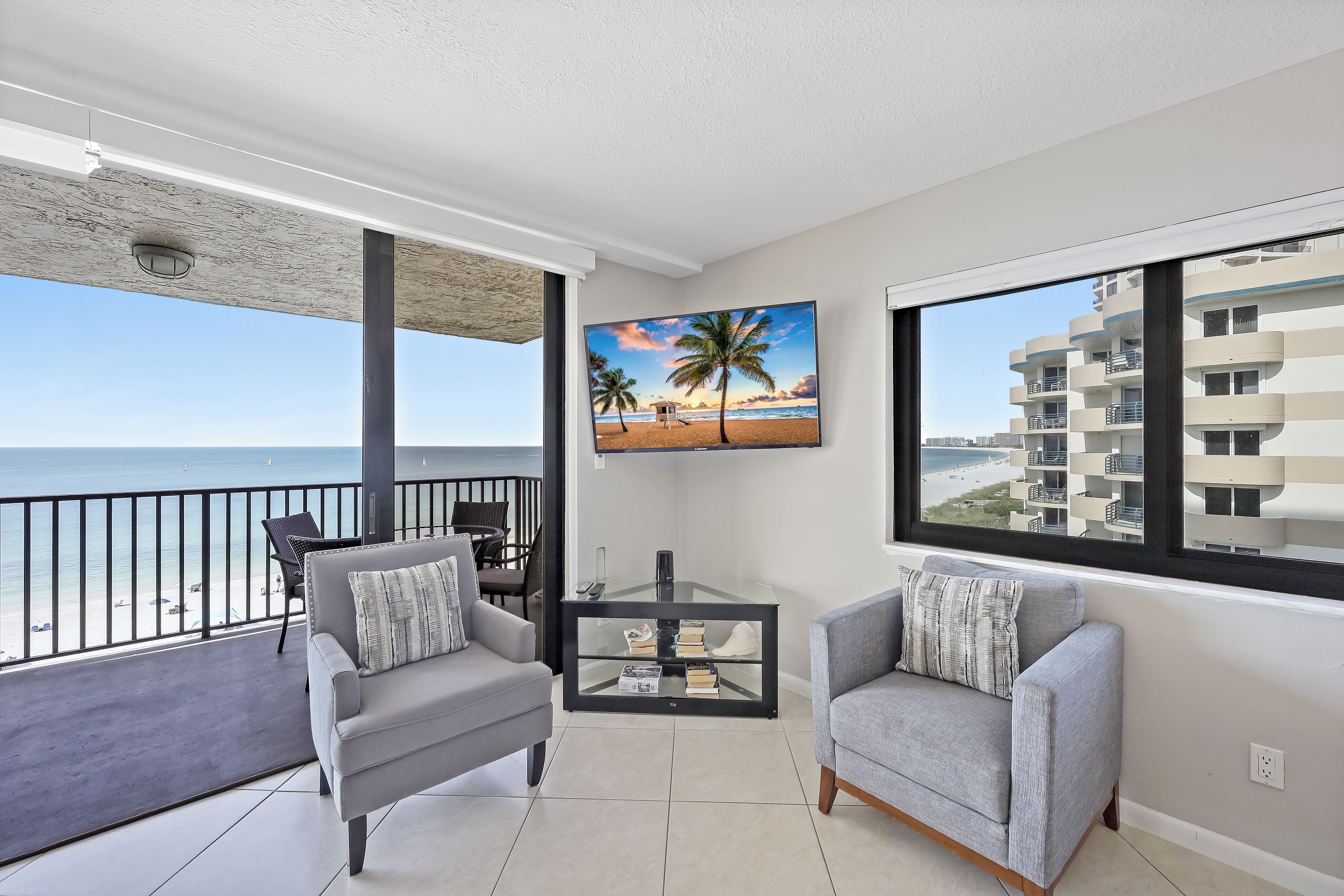 A balcony with a view of the beach, a TV mounted on the wall, and two grey sofas with cushions.
