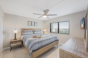 Une chambre à coucher comprenant un lit, des tables de chevet, un ventilateur de plafond, une fenêtre avec des stores et une vue sur l’extérieur.