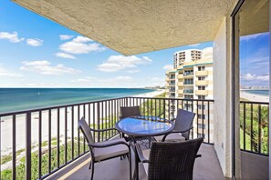 A balcony with a glass table and chairs, overlooking a beach and ocean. - Inviting Beachfront Condo | Pool, Spa & Views (Marco Island)