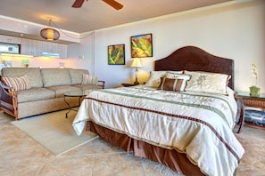 A bedroom with a large bed, a sofa, a dining table, and a kitchen area in the background.
