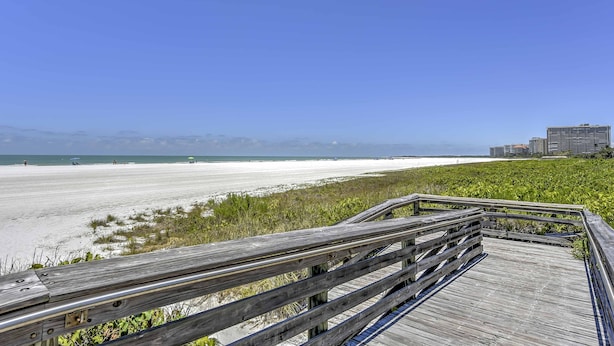 Una passerella di legno che conduce a una spiaggia, con vista libera sull'oceano e edifici in lontananza.