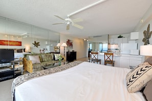 A modern hotel room with a large bed, a television, a dining area, and a kitchen.