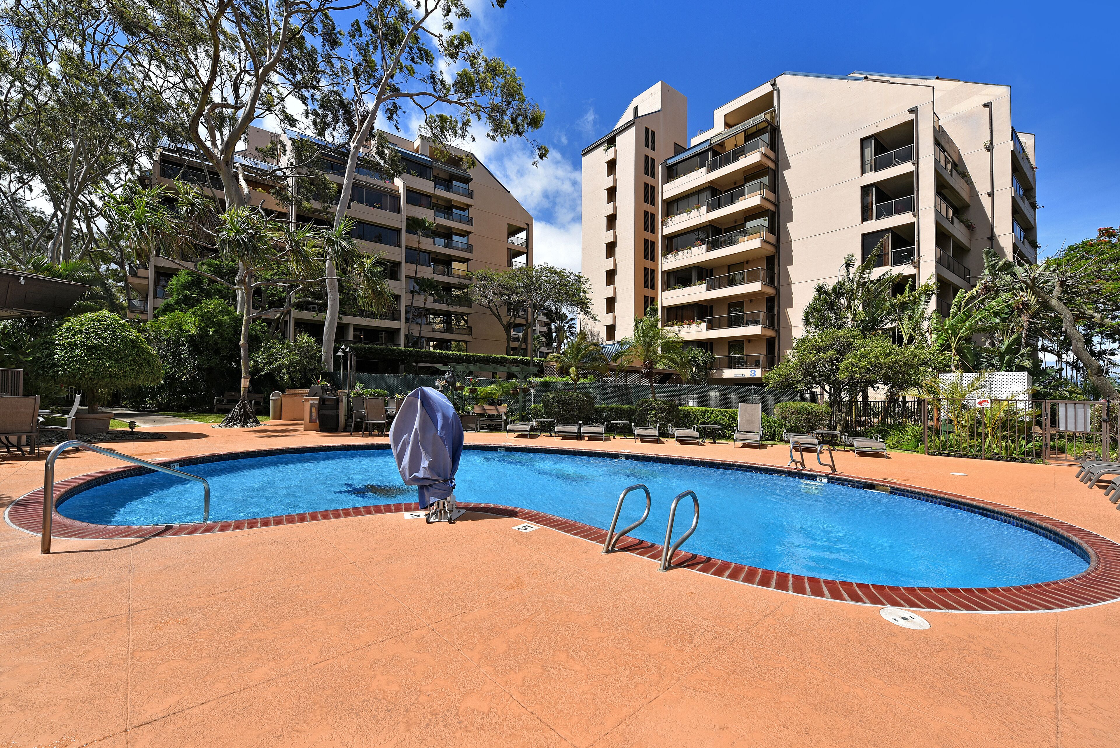 Una piscina con una silla cubierta, rodeada por una valla y contigua a un edificio de varios pisos con balcones.