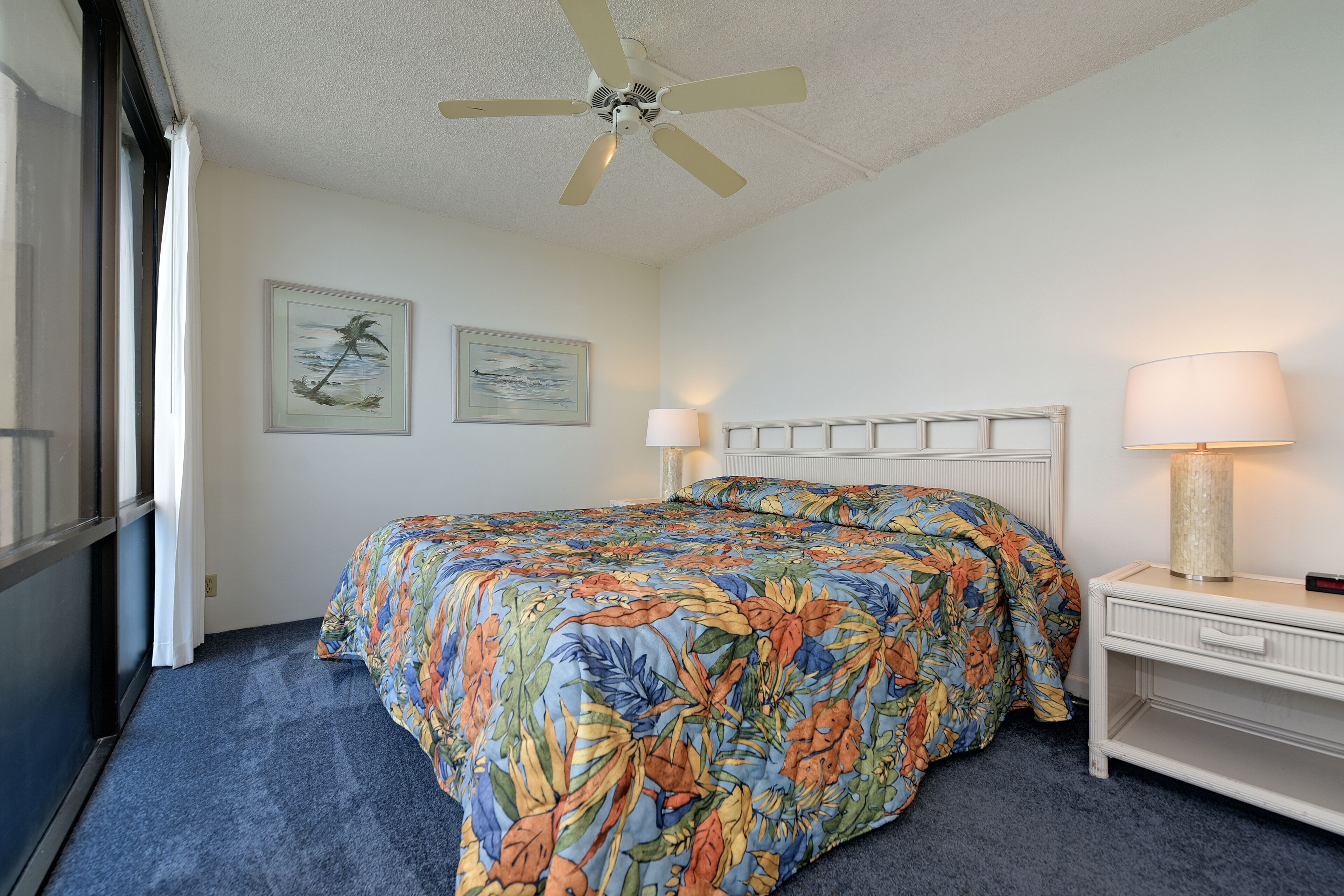 A hotel room with a bed, a ceiling fan, a nightstand with a lamp, and two framed pictures on the wall.