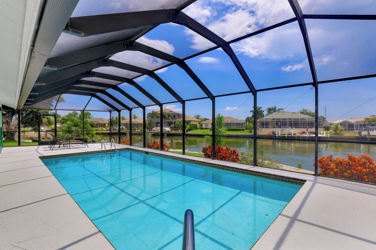 Une piscine dotée d’une structure en verre, entourée d’une terrasse et offrant une vue sur un lac et des maisons.