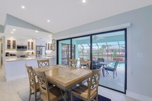 A modern kitchen with white cabinets and stainless steel appliances, a dining area with a wooden table and chairs, and a large sliding glass door leading to an outdoor patio with a pool and seating area. - Pet-Friendly Waterfront Home | Key Lot w/ View (Marco Island)