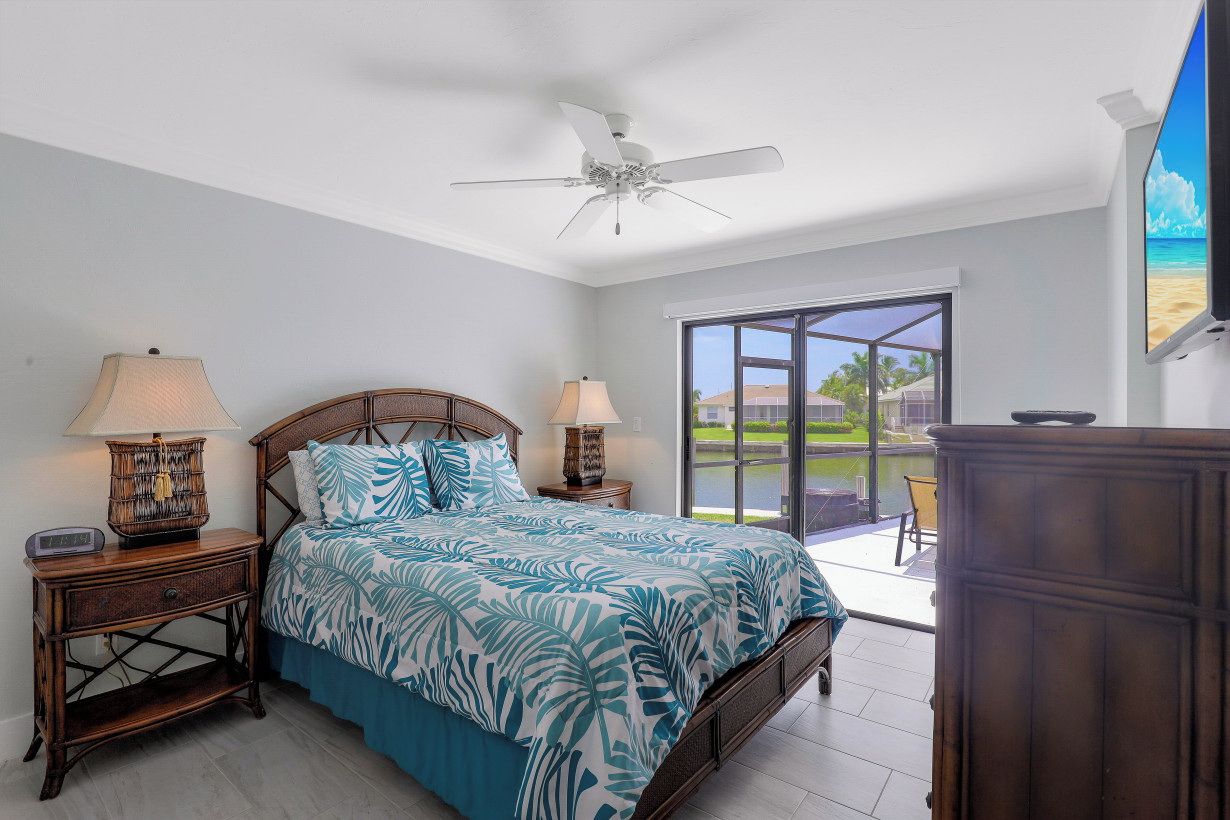 A bedroom with a bed, two bedside tables with lamps, a ceiling fan, and a view of the outdoors through a sliding glass door.