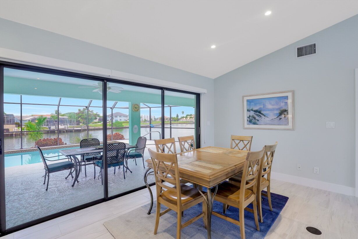 Un comedor con mesa y sillas de madera, ventanas amplias con vista a una piscina y follaje, y un cuadro en la pared.