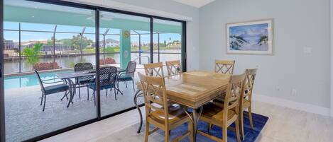 Un comedor con mesa y sillas de madera, ventanas amplias con vista a una piscina y follaje, y un cuadro en la pared.