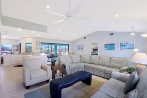 A spacious living area with a large L-shaped sofa, a blue bench, and a dining table with chairs. There is a ceiling fan, a chandelier, and artwork on the walls.