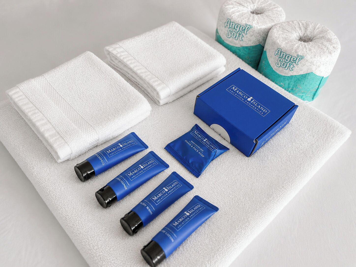 A set of toiletries including a shampoo bottle, hand wash, and soap, arranged on a white towel with a blue box and two towels.