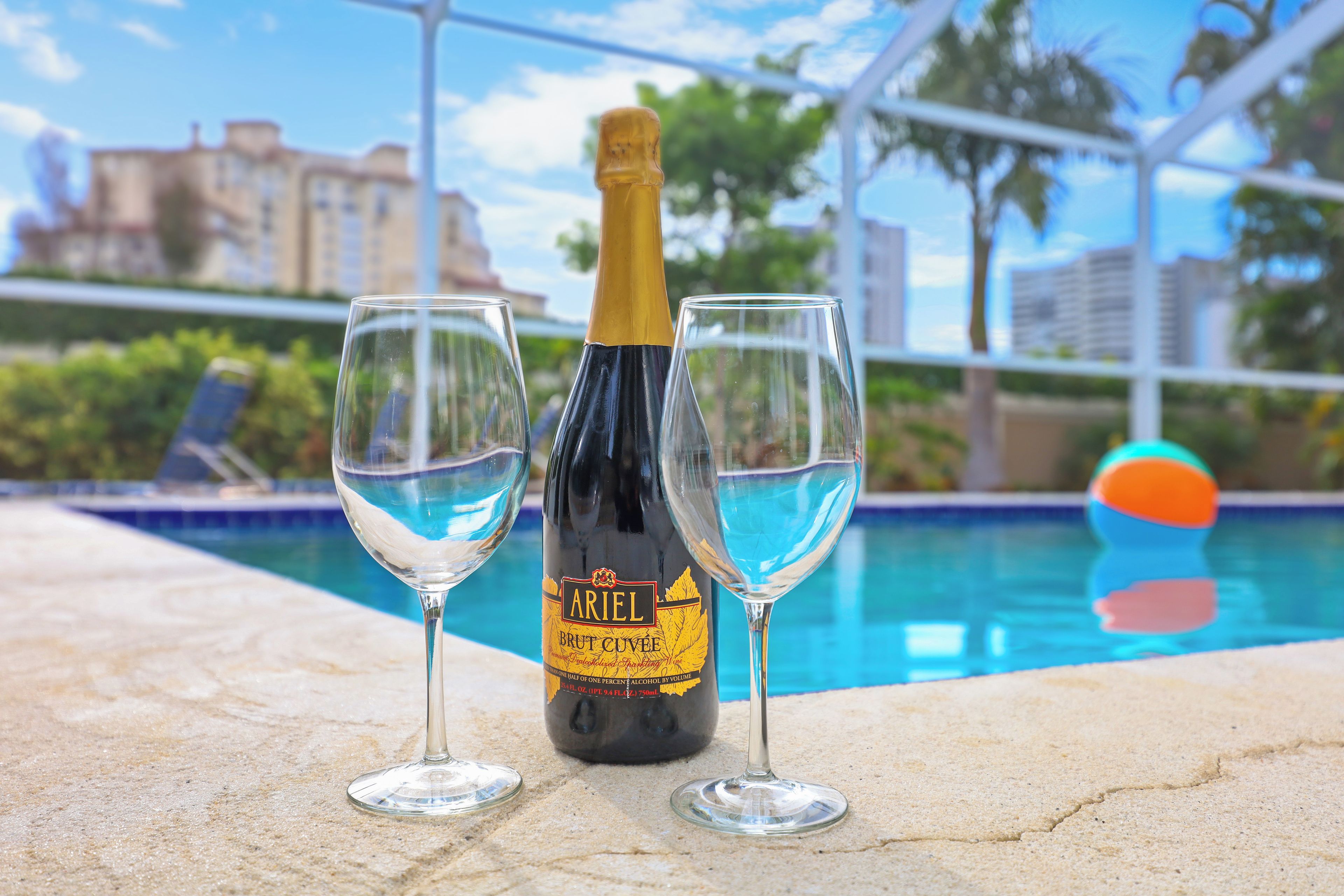 A bottle of Ariel Brut Cuvee champagne and two glasses on a poolside with a view of buildings and palm trees.