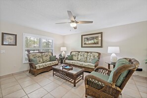 A living room with wicker furniture, a ceiling fan, and two windows with blinds.