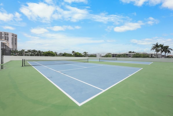 An outdoor tennis court with a net in the center, surrounded by a green surface and a blue playing area.