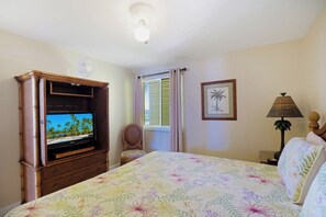 A bedroom with a bed, a television on a wooden cabinet, a window with curtains, a framed picture on the wall, and a lamp.