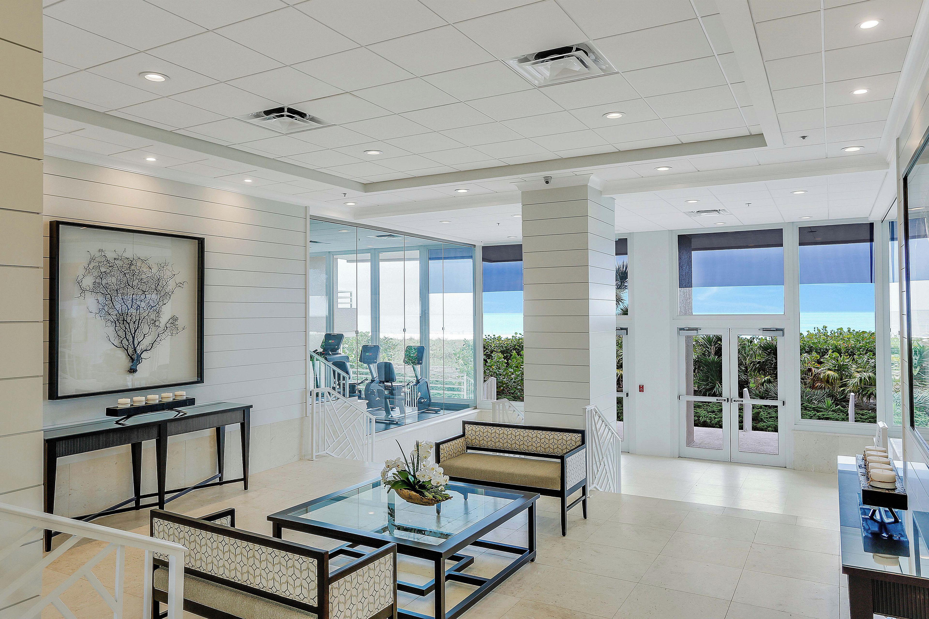 A modern hotel lobby with a glass wall offering a view of the outdoors, a seating area with a sofa and chairs, and a framed artwork on the wall.