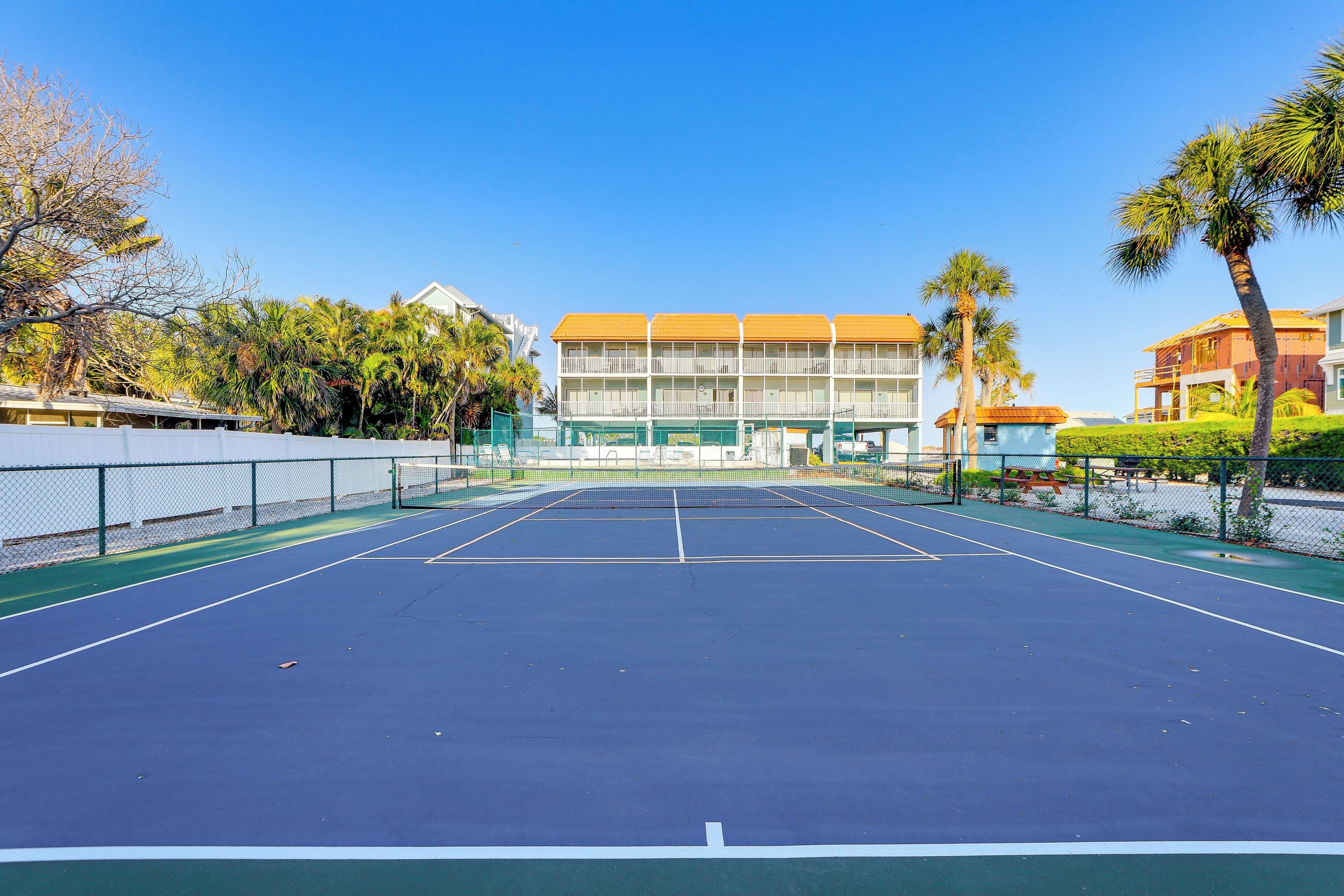 Un court de tennis avec un filet, entouré d’une clôture, et situé à côté d’un immeuble à plusieurs étages doté de balcons.
