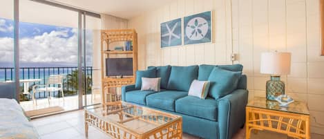 A room with a blue sofa, a glass coffee table, a wicker side table, and a view of the ocean through large windows.