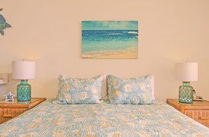 Une chambre à coucher avec un lit recouvert d’une couvre-lit à motifs bleus et blancs, deux oreillers assortis et deux tables de chevet avec des lampes. Un tableau représentant une scène de plage est accroché au mur, au-dessus du lit.