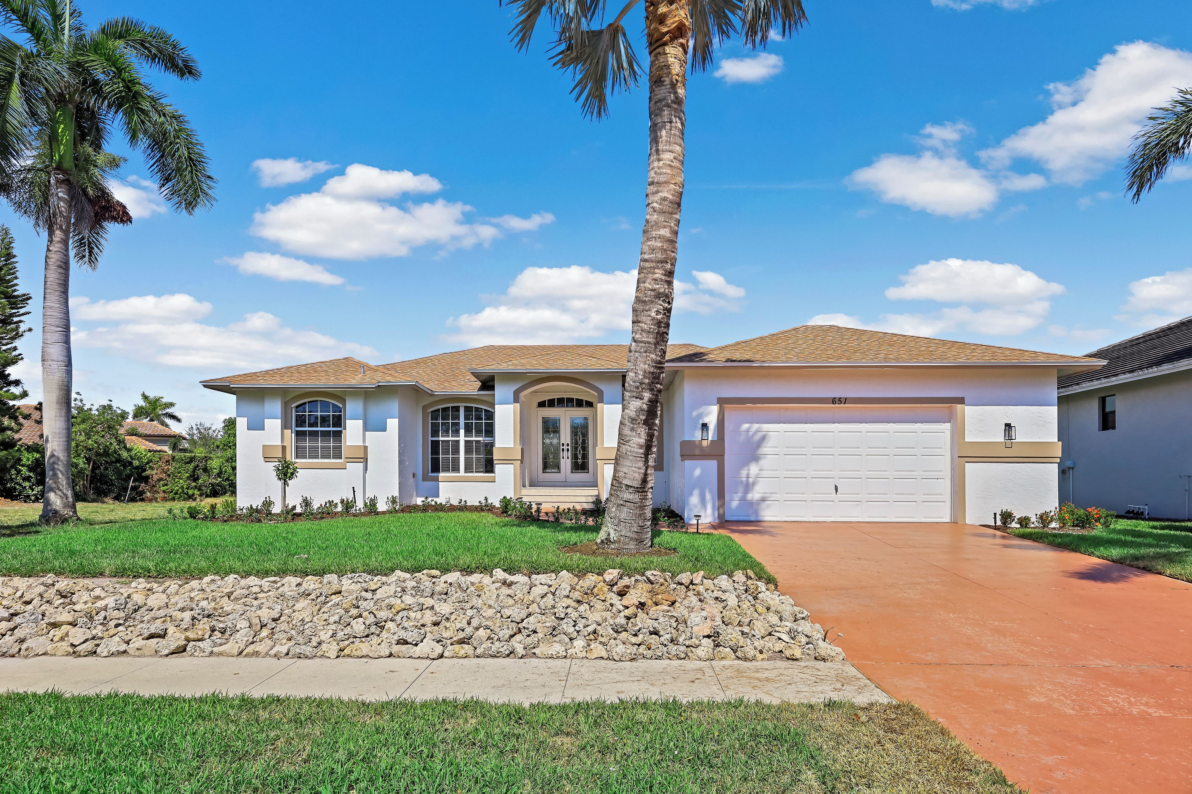 A single-story house with a garage, surrounded by a well-maintained lawn and palm trees.