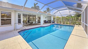 A covered swimming pool with a clear view of the surrounding area, including a patio with chairs and a table, and a view of palm trees and a building in the background. - Inviting and Quiet Home | Pool & Steps to Beach (Marco Island)