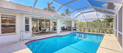A covered swimming pool with a clear view of the surrounding area, including a patio with chairs and a table, and a view of palm trees and a building in the background.