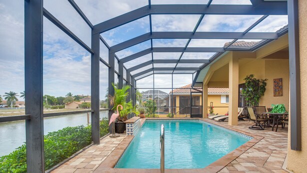 An indoor swimming pool with a glass ceiling, surrounded by a patio area with chairs and a table, and a view of a body of water and buildings in the background. - Upscale Marco Island Villa w/ Outdoor Bar + Pool! (Marco Island)
