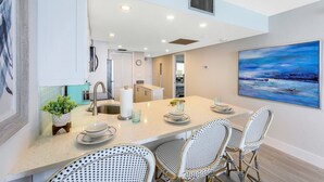 A modern kitchen with a large island, white cabinetry, and a dining area with wicker chairs. - Charming Beachfront Condo | Pool, Spa & Views (Marco Island)