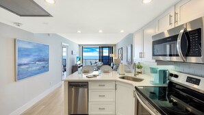 A modern kitchen with white cabinetry, stainless steel appliances, and a view to the outside. - Charming Beachfront Condo | Pool, Spa & Views (Marco Island)