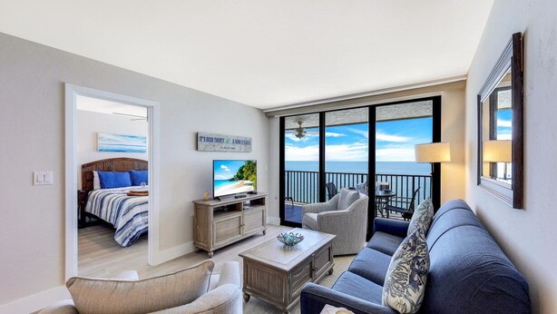 A modern living room with a sofa, chairs, a coffee table, and a television. A balcony with a view of the ocean is visible through large windows. - Charming Beachfront Condo | Pool, Spa & Views (Marco Island)