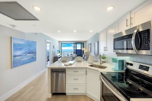 A modern kitchen with white cabinetry, stainless steel appliances, and a view to the outside. - Charming Beachfront Condo | Pool, Spa & Views (Marco Island)