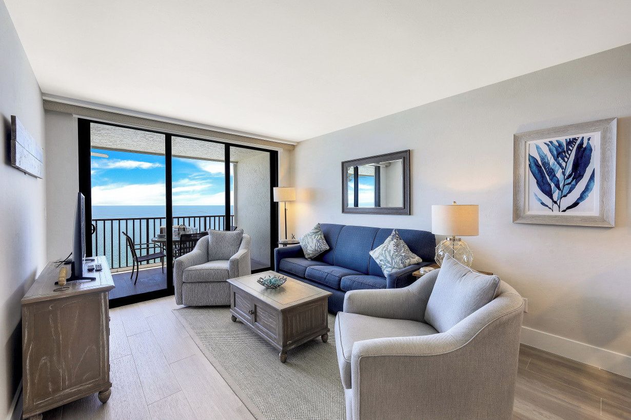 A modern living room with a blue sofa, two armchairs, a coffee table, and a TV. There is a balcony with a view of the sea.