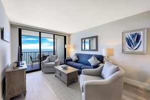 A modern living room with a blue sofa, two armchairs, a coffee table, and a TV. There is a balcony with a view of the sea.