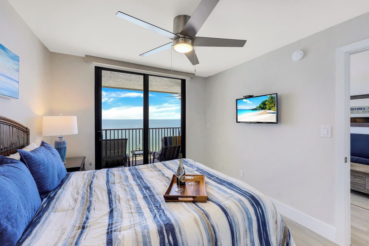 Hotelkamer met een bed, een plafondventilator, een televisie, een balkon met uitzicht op de oceaan en een dienblad met een glas en een fles.