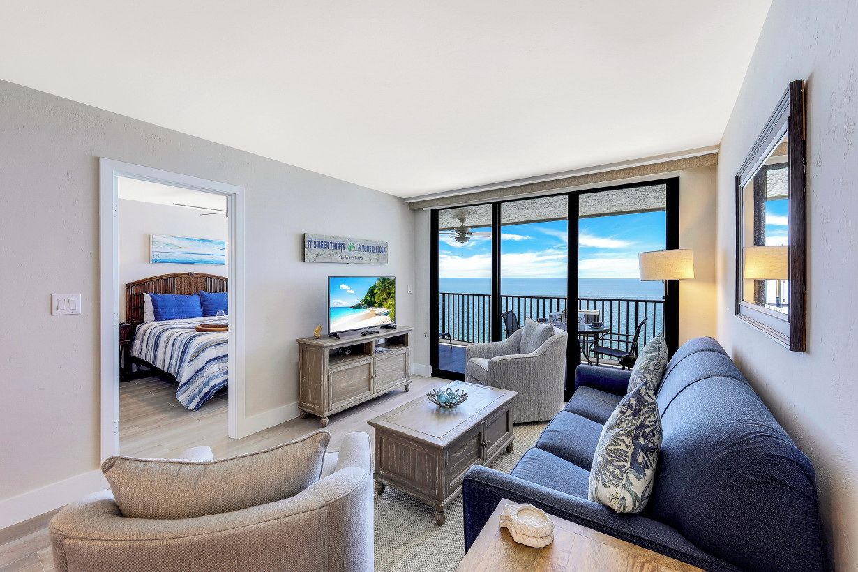 A modern living room with a sofa, chairs, a coffee table, and a television. A balcony with a view of the ocean is visible through large windows.