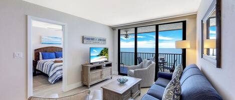 A modern living room with a sofa, chairs, a coffee table, and a television. A balcony with a view of the ocean is visible through large windows.