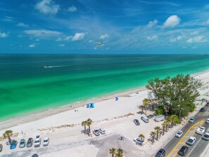 A beach with clear turquoise waters, a parking area with several cars, and a few people walking along the shore.