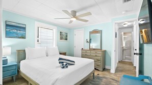 Un dormitorio con cama, cómoda, espejo, ventilador de techo y un baño visible a través de una puerta abierta.