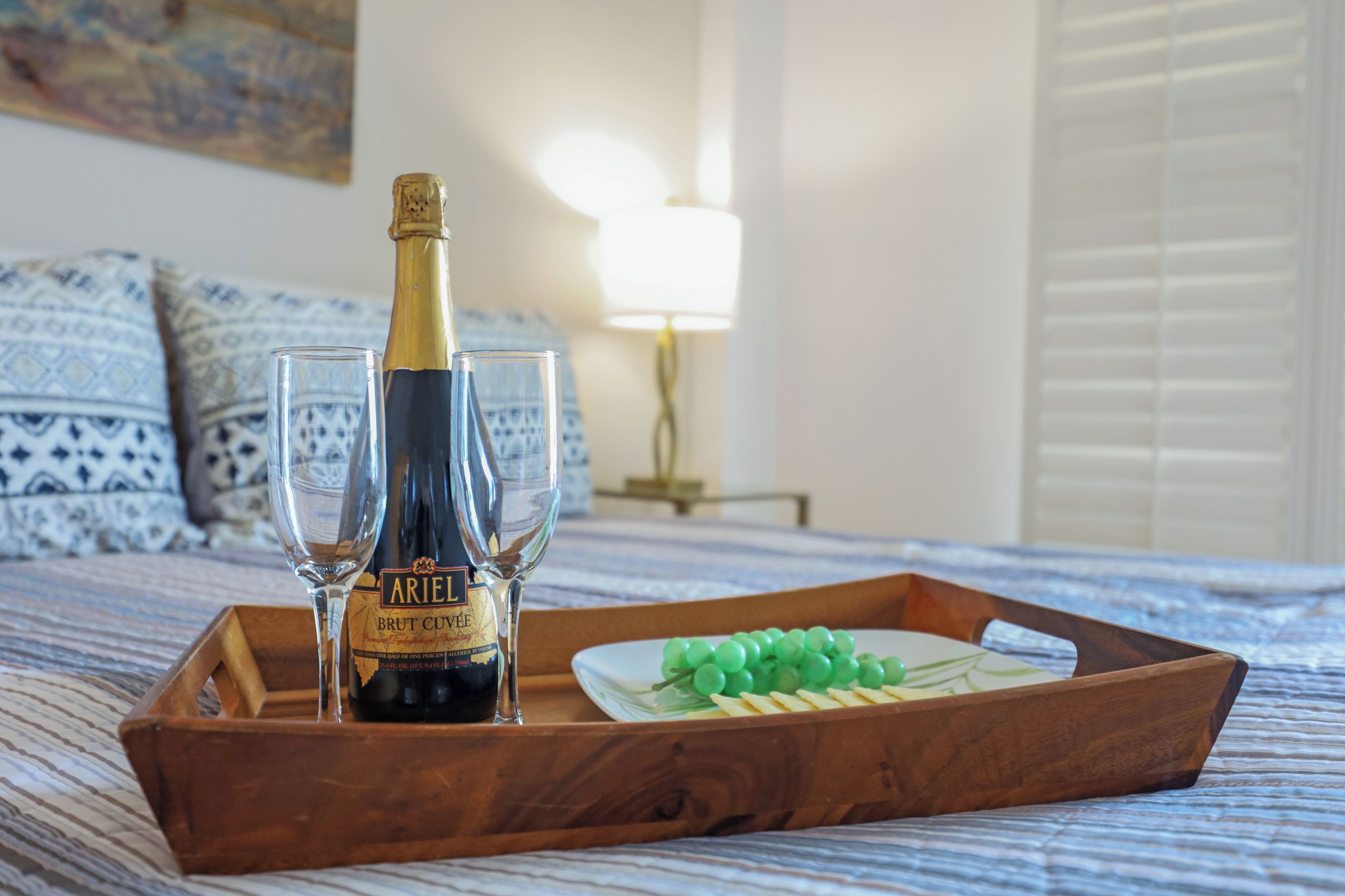 A wooden tray with a bottle of Brut Cuvée champagne, two glasses, and a plate of green grapes on a bed.