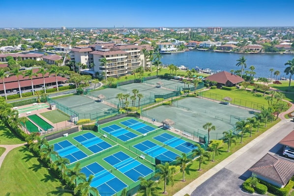 A large sports complex with multiple tennis courts, surrounded by residential buildings and a body of water.